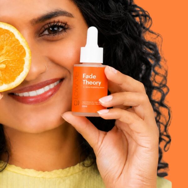 Woman holding Fade Theory 2% alpha arbutin serum beside her face, with a slice of orange, on an orange background