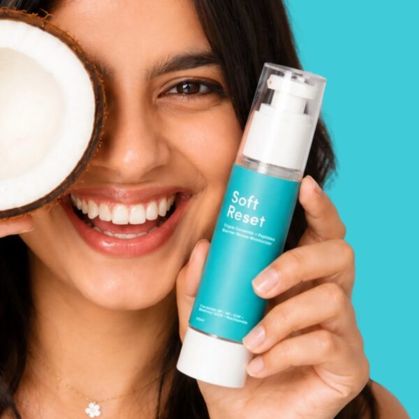 Woman holding Soft Reset barrier repair moisturiser beside her face, with half a coconut, on a teal background